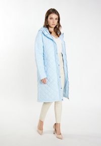 Cappotto lungo trapuntato blu chiaro con cappuccio, caratterizzato da chiusura a bottoni e tasche laterali, abbinato a pantaloni slim color crema e tacchi beige.