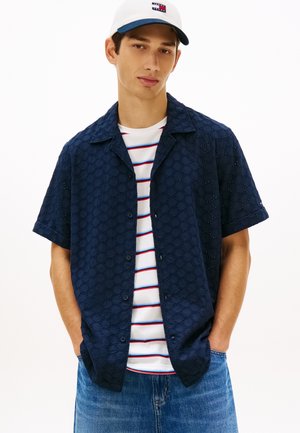 Jonge man met een witte pet, een marineblauw tekstured overhemd met korte mouwen over een wit T-shirt met strepen, en een blauwe spijkerbroek met handen in de zakken.