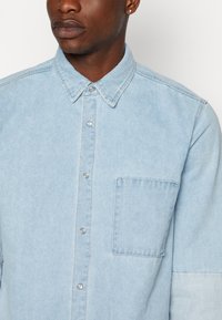 Camicia in denim azzurro chiaro con chiusura frontale a bottoni, colletto a punta e una tasca sul petto. Presenta cuciture in vista e bottoni argentati.