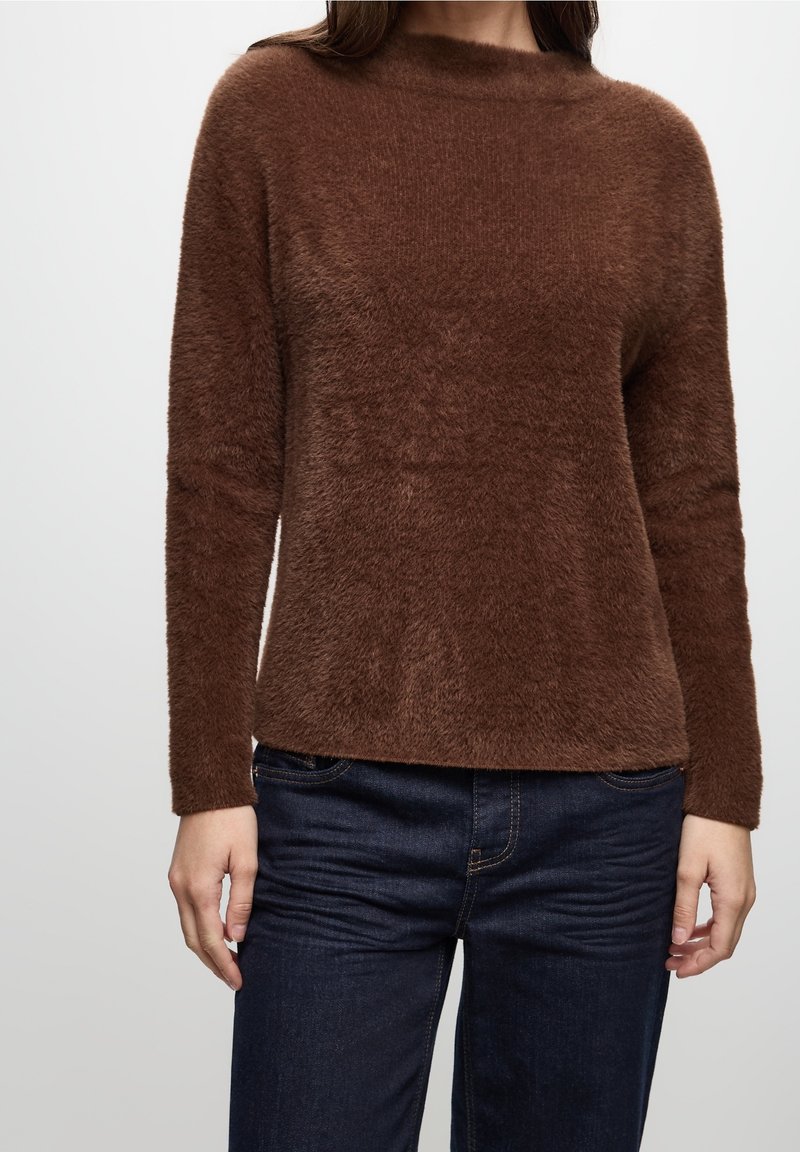 Femme portant un pull marron duveteux à manches longues associé à un jean bleu foncé, debout devant un fond clair uni.