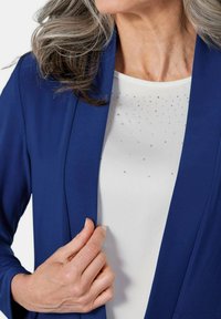 Marineblauer Blazer mit tailliertem Schnitt, kombiniert mit einem weißen Oberteil, das mit verstreuten Strasssteinen verziert ist. Glattes Material und elegante Revers.