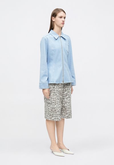 Ανοιχτό μπλε τζιν μπουφάν με φερμουάρ και μια μπροστινή τσέπη, σε συνδυασμό με γκρι cargo σορτς με λεοπάρ μοτίβο και λευκά παπούτσια τύπου slip-on.