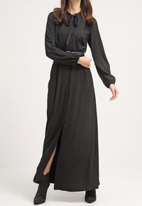 Robe maxi noire avec une fermeture à boutons sur le devant, des manches longues, un col noué et une fente latérale, confectionnée dans un tissu lisse. Soulignée par une ceinture.