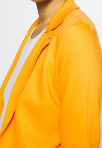 Blazer arancione con una texture liscia, dotato di colletto a revers e una spilletta decorativa sul davanti. La camicia bianca sottostante è parzialmente visibile.