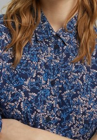 Chemise bleu marine à boutons avec un motif floral bleu et rose, dotée d'un col et d'un tissu texturé.