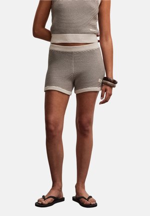 Person iført beige teksturerede strikkede shorts og matchende ærmeløs top, tilbehør med to armbånd og sorte klip-klappere.