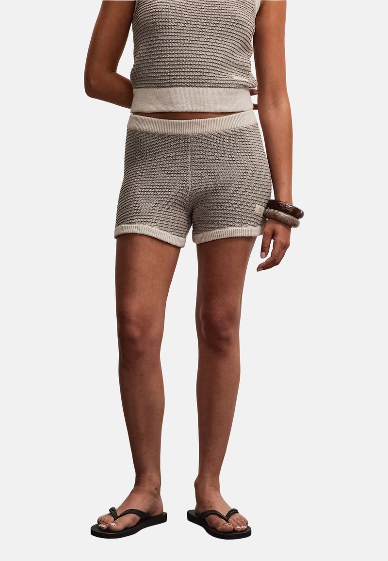 Personne portant un short en maille beige texturé et un top sans manches assorti, accessoirisés de deux bracelets et de sandales tongs noires.