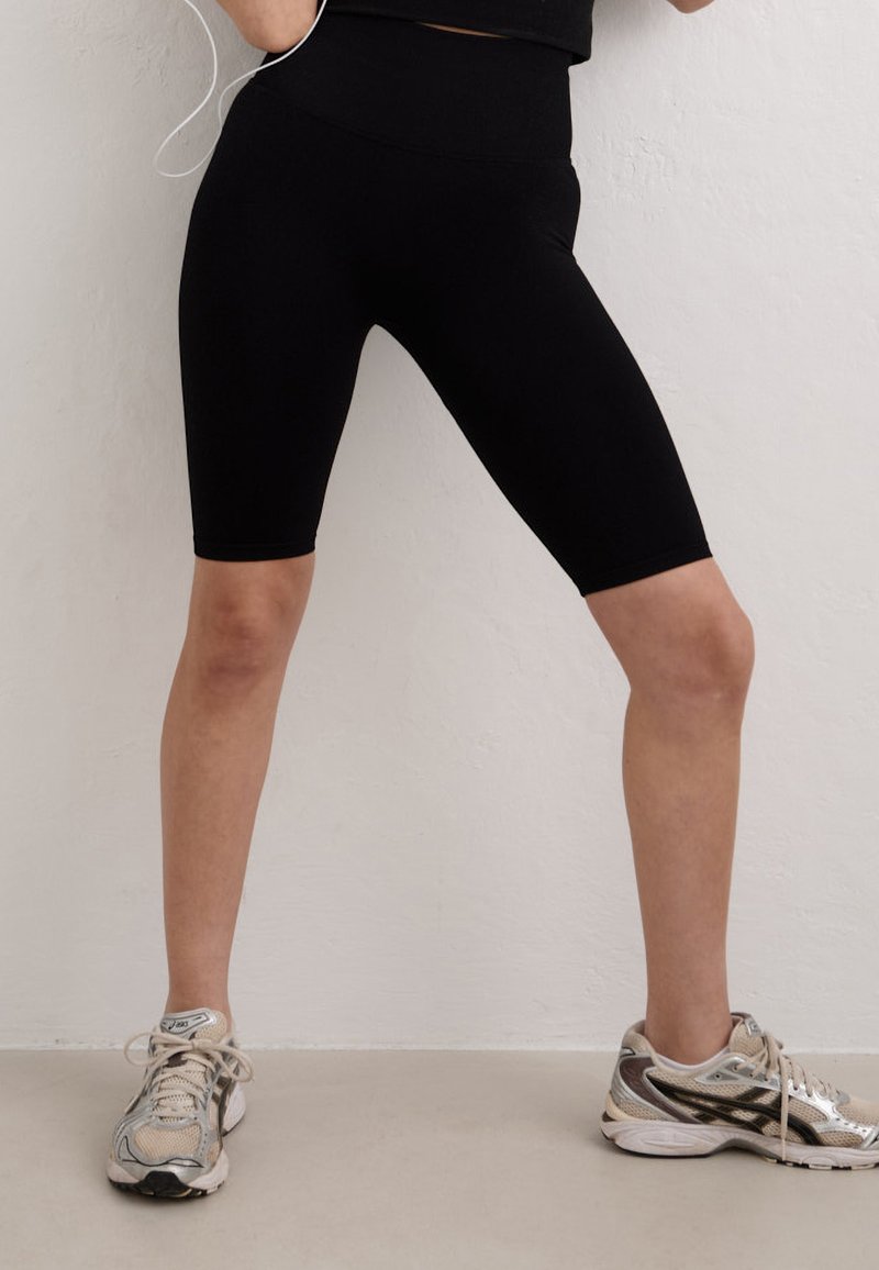 Personne portant des leggings noirs jusqu'aux genoux et des chaussures de course beige, debout jambes écartées contre un mur clair.