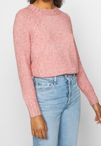 Pull en maille rose clair avec poignets côtelés et encolure ronde, présentant une coupe décontractée et un tissu texturé, assorti à un jean bleu clair.