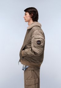 Khaki kurtka bomber z kołnierzem z sztucznego futra, z zamkiem z przodu, bocznymi kieszeniami oraz naszywką na rękawie, w połączeniu z beżowymi spodniami cargo.