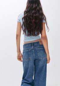 Högmidjade blå denimjeans med avslappnad passform, som har två bakfickor och ett beige midjeband, sedd bakifrån.