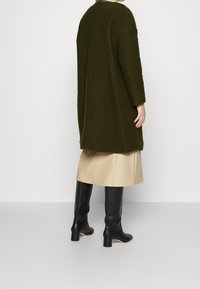 Cappotto lungo verde oliva, lavorato con una vestibilità ampia, spalle scese e maniche lunghe, abbinato a una gonna beige e stivali neri al ginocchio.