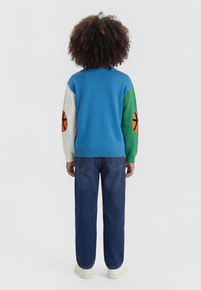 Παιδί με σγουρά μαλλιά στέκεται με την πλάτη, φοράει μπλε πουλόβερ με λευκά και πράσινα μανίκια με σχέδια καλαθοσφαίρισης και μπλε τζιν.