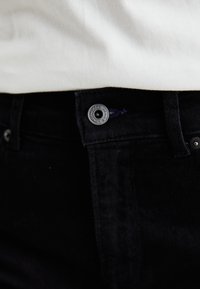 Los pantalones de mezclilla negra presentan un diseño ajustado con un estilo clásico de cinco bolsillos. La cinturilla incluye un botón de metal y un acento de costura azul.