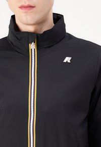 Čierna mikina na zips s vysokým golierom, ktorý má farebný akcent zipsu s pruhmi žltej, bielej a modrej farby, značkový logotyp na hrudi.