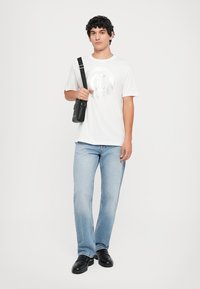 En ung mann iført en hvit grafisk t-skjorte, lyseblå jeans, svarte sko og en svart crossover-veske, står mot en ensfarget bakgrunn.