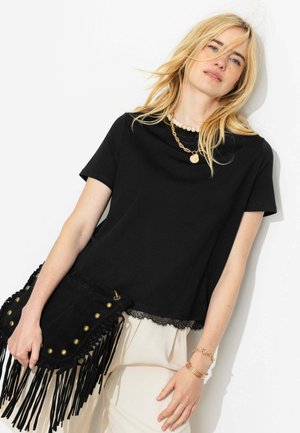 Femme blonde portant un top noir à manches courtes avec bordure en dentelle, un pantalon crème, des bijoux en or et tenant un sac à franges noir avec des clous en laiton.