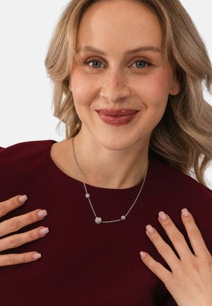 Lächelnde Frau mit blondem Haar und Sommersprossen, die ein burgunderrotes Oberteil und eine silberne Halskette mit kleinen dekorativen Perlen trägt, Hände in der Nähe des Schlüsselbeins.