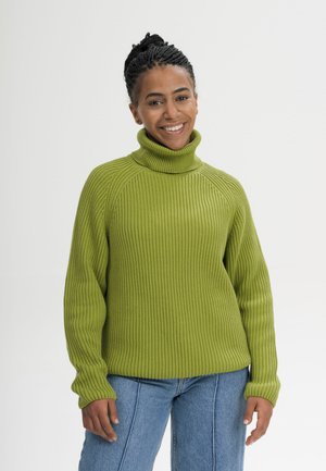 Vrouw met gevlochten haar die glimlacht, draagt een groene geribde coltrui en blauwe jeans, staande tegen een effen lichte achtergrond.