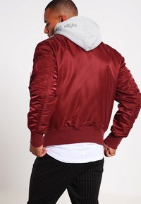 Giacca bomber bordeaux con finitura lucida, polsini e orlo a coste. Presenta un cappuccio grigio che spunta da sotto, con dettagli di tasche sulle maniche.