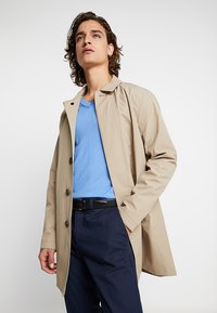 Abrigo impermeable beige con botón en el frente, amplias solapas y mangas largas. Combinado con una camiseta azul de escote en V y pantalones azul marino texturizados.