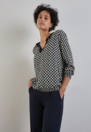 Gemusterte Bluse in Schwarz und Creme mit V-Ausschnitt und langen Ärmeln, mit elastischem Saum. Kombiniert mit dunklen Hosen.