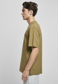 Tricou din bumbac de culoare verde măsliniu, cu mâneci scurte, guler rotund și o croială lejeră, prezentat din lateral, asortat cu blugi din denim deschis.