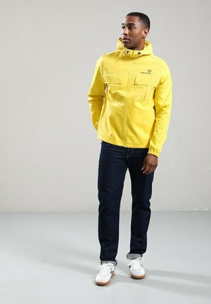 Uomo che indossa una giacca gialla con cappuccio, jeans scuri e scarpe da ginnastica bianche, in piedi su uno sfondo grigio chiaro uniforme.