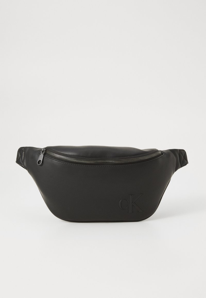 Borsa vita in pelle nera con chiusura a zip e logo "cK" impresso sul davanti, mostrata su uno sfondo bianco neutro.