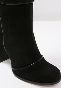 Svart mocka ankelsko med rund tå, sid sömdetaljer och en glänsande svart accent i mitten. Den har en kraftig klack.