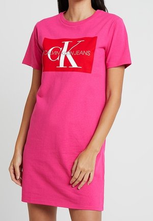 Różowa bawełniana sukienka t-shirtowa z dużą czerwoną grafiką z napisem "CALVIN KLEIN JEANS" oraz wyraźnym białym logo "CK" z przodu.