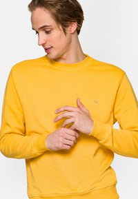 Żółty sweatshirts z okrągłym dekoltem i długimi rękawami, o miękkiej fakturze, z małym niebieskim logo na piersi. Model dostosowuje mankiety.