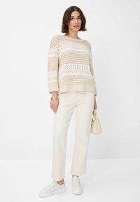 Maglione beige lavorato a maglia con motivo a pizzo, righe bianche, vestibilità rilassata, abbinato a pantaloni avorio e sneakers bianche. Borsa minimalista in mano.
