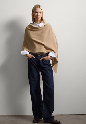Un abrigo beige de material suave se drapea sobre una camisa blanca con cuello, combinado con unos vaqueros de pierna ancha azul oscuro y zapatos marrones. Diseño simple y limpio.