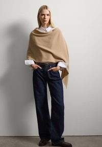 Un wrap beige realizzato in materiale morbido si adagia su una camicia bianca con colletto, abbinato a jeans a gamba larga blu scuro e scarpe marroni. Design semplice e pulito.