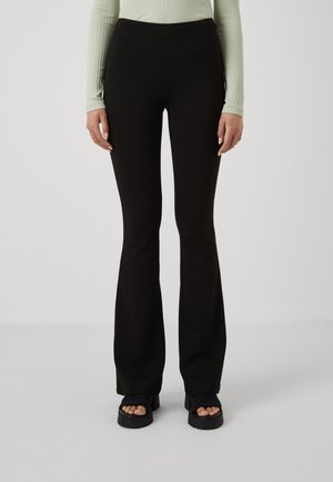 Femme portant un pantalon noir évasé, un haut à manches longues côtelé vert clair et des sandales à plateforme noires, debout sur un fond uni.