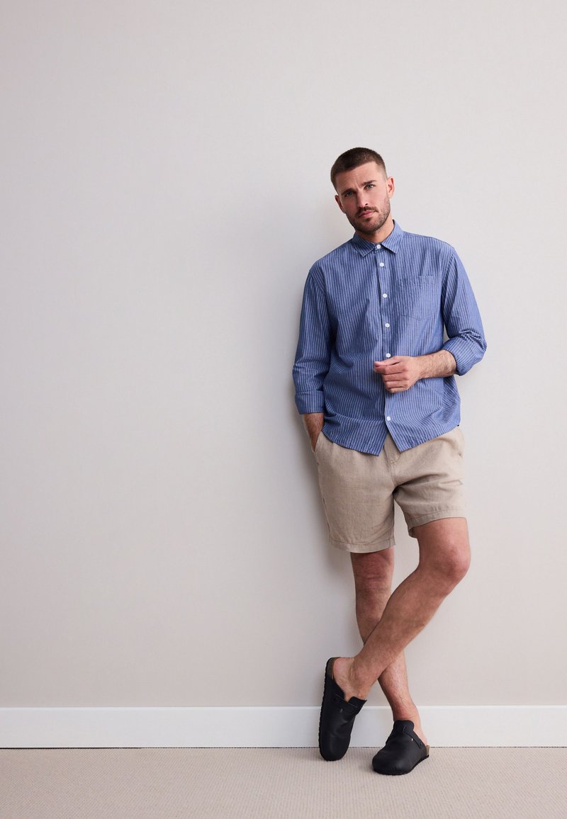 Camicia a righe bianche e blu con bottoni, pantaloncini beige in lino, scarpe nere senza lacci. Modello appoggiato a una parete, braccia rilassate.