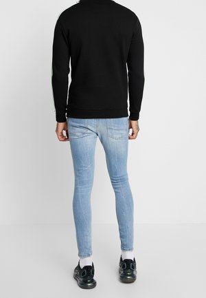 Mężczyzna w jasnoniebieskich obcisłych dżinsach, czarnej kurtce z zielonymi i białymi paskami, białych skarpetkach i czarnych trampkach stoi oparty o białą ścianę.