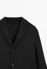 Cappotto di lana nera con collo a scarico e due bottoni frontali. Il tessuto ha una finitura morbida e texturizzata senza motivi visibili.