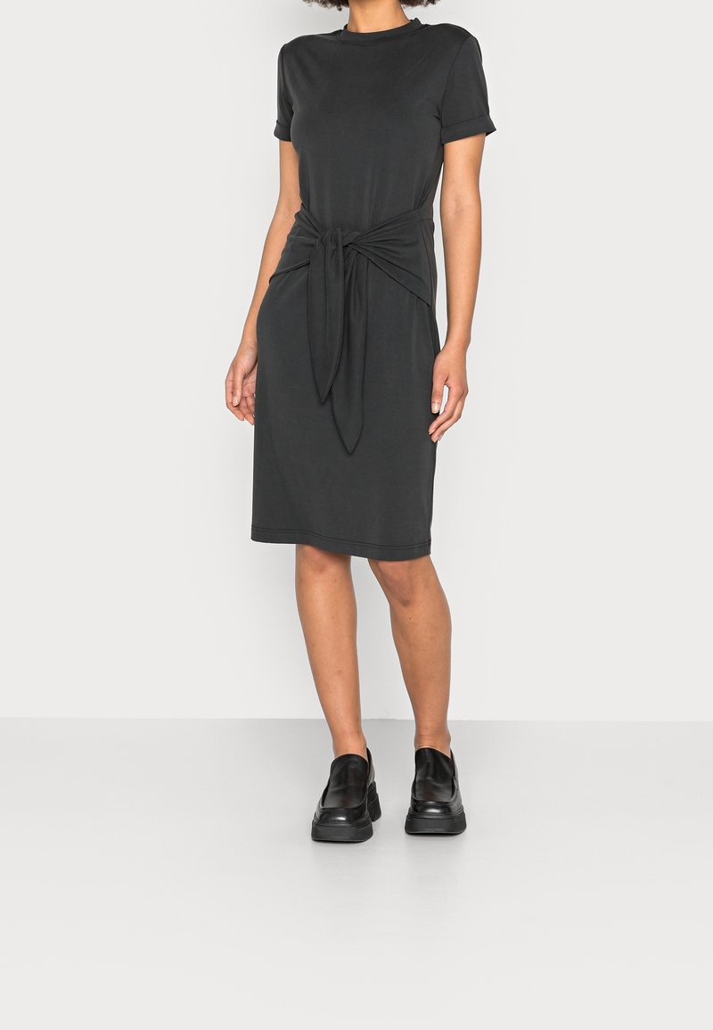 Femme portant une robe noire courte à manches courtes avec un détail de nœud à l'avant, associée à des chaussures noires à plateforme et sans lacets.
