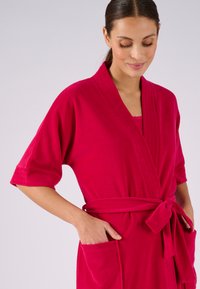 Fuchsia badjas met korte mouwen, strikceintuur en twee voorzakken; gemaakt van zachte, gestructureerde stof met subtiele verticale naden.