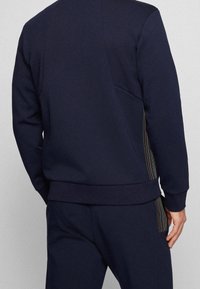 Person i en marinblå träningsoverall med vertikala flerfärgade ränder på sidorna, visad bakifrån mot en enkel bakgrund.