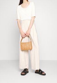 Bílá volná tričko, béžové široké kalhoty, černé sandály na řemínky a malá tkaná kabelka v hnědé barvě se hladkým povrchem.