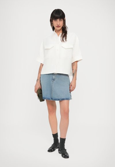 Λευκό πουκάμισο με κοντά μανίκια και δύο τσέπες στο στήθος, συνδυασμένο με φούστα από ανοιχτό μπλε τζιν. Μαύρα παπούτσια με λευκές λεπτομέρειες, σκούρες κάλτσες και πράσινο τσαντάκι χειρός.