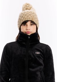 Zwarte fleece zip-up jas met hoge kraag en een logo patch, gecombineerd met een beige gebreide beanie met een pompon bovenop.