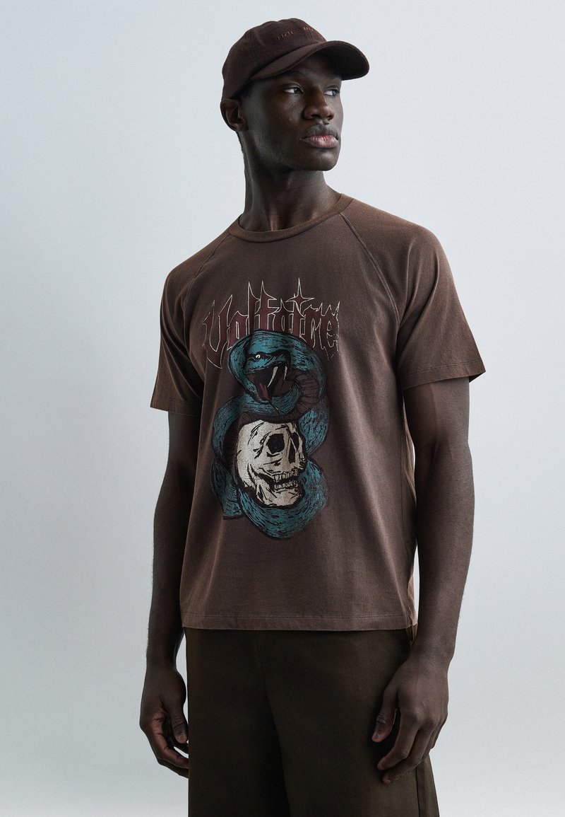 Άνδρας που φορά καφέ καπέλο και καφέ μπλούζα με σχέδιο μπλε φιδιού τυλιγμένου γύρω από κρανίο και το κείμενο «Voltfire», κοιτάζοντας προς τα πλάγια σε απλό φόντο.