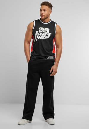 Schwarzes sportliches Tanktop mit weißen und roten Akzenten; verfügt über einen auffälligen Grafikdruck. Kombiniert mit schwarzen Jogginghosen und weißen Sneakers.