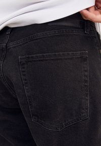 Närbild av svarta jeans på en person, som visar bakfickans sömnad och midjebandet med en vit skjorta delvis instoppad.