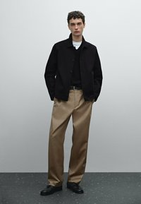 Giacca nera con bottoni sopra un maglione nero, abbinata a pantaloni larghi beige e scarpe nere con lacci. Design semplice e su misura.