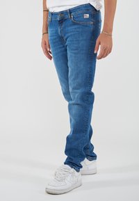 Jeans in denim blu con taglio dritto, cinque tasche e classico bottone a pressione. Indossati con sneakers bianche da ginnastica.